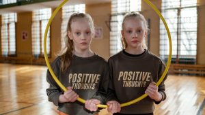 Twin girls in gym holding a hula hoop, wearing 'Positive Thoughts' sweatshirts.